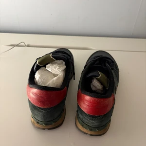 Valentino Garavani Rockrunner sneakers - Snygga Valentino Garavani Rockrunner sneakers i mörkgrönt och svart skinn och mocka, med röda detaljer på hälen och klassisk gummisula. Skorna har snörning och cool camouflagemönstrad design som sticker ut. Perfekta för dig som vill ha något unikt och trendigt.