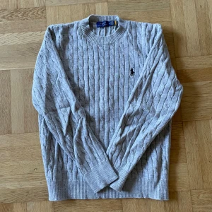 Polo Ralph Lauren tröja - Säljer en grå kabelstickad tröja från Polo Ralph Lauren i storlek M. Tröjan har rund hals, lång ärm och klassisk broderad logga på bröstet. Tillverkad i mjuk bomull, perfekt för dig som gillar stilrena och tidlösa plagg.