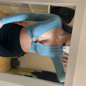 Ljusblå omlottopp med ringdetalj - Trendig ljusblå omlottopp från PLT med lång ärm, croppad passform och snygg ringdetalj framtill. Toppen har djup v-ringning, öppen rygg och knytband bak för extra cool look. Perfekt för dig som vill sticka ut med en unik och stilren topp.