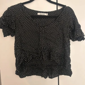 Svart prickig blus från NA-KD - Supersöt svart blus från NA-KD med vita prickar, volangdetaljer och knyt framtill. Blusen har korta ärmar med volang och en lätt croppad passform. Perfekt för dig som gillar en lekfull och trendig stil.
