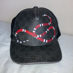 Gucci svart keps med ormprint - Snygg svart keps från Gucci med mesh på sidorna och bak, samt justerbar rem med röd och blå detalj. Framtill pryds kepsen av en stor röd, vit och svart orm och diskret GG-mönster över hela tyget. Materialet är bomull och polyester.