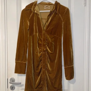 Guldbrun sammetsklänning Urban Outfitters - Supersnygg kort sammetsklänning i guldbrun ton från Urban Outfitters. Klänningen har långa ärmar, breda manschetter med knappar, krage och knappar hela vägen framtill. Snygg rynkning längs knappraden ger en cool vintagevibe. Perfekt för dig som gillar unika detaljer!