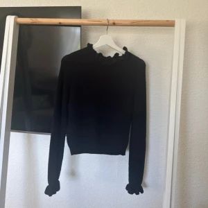 Svart stickad tröja med volangkrage - Säljer en svart stickad tröja med lång ärm och volangdetaljer vid både halsringning och ärmslut. Tröjan har en klassisk passform. 