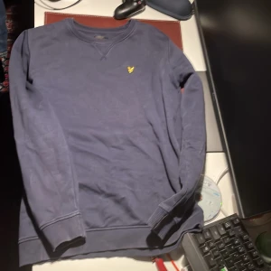 Blå sweatshirt från Lyle & Scott - Mörkblå sweatshirt från Lyle & Scott med klassisk rund hals och gul logga broderad på bröstet. Tröjan har långa ärmar och ribbade muddar vid ärmslut och nederkant. Perfekt för en chill och stilren look.