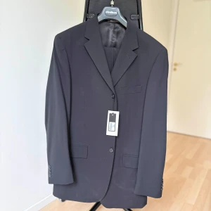 Ny svart kostym från Medi – Made in Italy, stl 52 - Säljer en helt ny svart kostym från Medi, Made in Italy. Kostymen är i klassisk modell med två knappar, ficklock och innerfoder i satin. Byxor med pressveck ingår.  Storlek: 52 (motsvarar ca L/50–52 EU)  Färg: Svart (kod 9905)  Material: Ullblandning (Made in Italy-kvalitet)  Nypris: 4500 kr  Skick: Helt ny med tags kvar  Passar både till arbete, fest och bröllop.