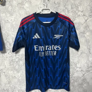 Arsenal Rice #41 blå Adidas tröja S - Snygg Arsenal fotbollströja från Adidas i storlek S, med Rice och nummer 41 på ryggen. Tröjan är marinblå med blixtmönster i blått, röda ränder på axlarna och vita tryck. Slim fit i lätt polyester, perfekt för match eller träning.