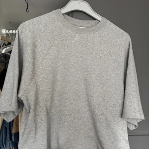 Grå sweatshirt med kort ärm - En basic grå sweatshirt i mjuk bomull med rund halsringning och korta ärmar. Perfekt för en avslappnad stil och enkel att matcha med jeans eller shorts. Klassisk och stilren design utan tryck eller mönster.