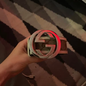 Limiterat gucci bälte  - Säljer ett klassiskt Gucci-bälte i beige och brunt med GG-monogram längs hela bandet. Bältet har en stor, rund spänne i silverfärgad metall och detaljer i ljust skinn. Perfekt accessoar för att lyfta din outfit med en lyxig touch.