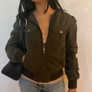 Olivgrön jacka - Snygg olivgrön jacka med luva och dragkedja framtill. Jackan har två bröstfickor med knappar, ribbade muddar och midja samt detaljerade sömmar. Perfekt för dig som vill ha en chill och stilren look till vardags.