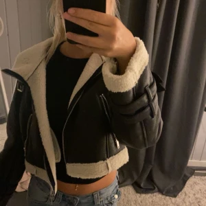 Svart teddyjacka från Boohoo - Säljer en svart croppad skinn teddyjacka från Boohoo med vit fuskpäls på insidan och vid kanterna. Jackan har dragkedja, långa ärmar och coola detaljer som spännen på ärmarna. Perfekt för dig som vill ha en trendig och mysig look i höst och vinter.