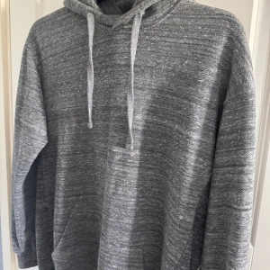 Grå hoodie från Vaillent - Säljer en gråmelerad hoodie från Vaillent med huva och snörning. Tröjan har en klassisk känguruficka framtill och är långärmad. Perfekt för chill dagar eller när du vill ha något bekvämt och stilrent.