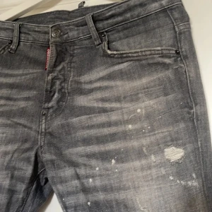 Grå Dsquared2 jeans - Säljer ett par grå jeans från Dsquared2 med coola slitningar och färgstänk för en edgy vibe. Jeansen har raka ben, klassisk femficksdesign och snygg Dsquared2-patch bak i midjan. Perfekta för dig som gillar streetstyle och vill sticka ut.