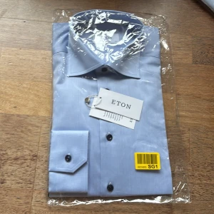 Ljusblå skjorta från Eton i bomull - Snygg ljusblå skjorta från Eton med klassisk krage och mörkblå knappar. Skjortan är tillverkad i bomull och har en stilren look som passar perfekt till jobbet eller en date. Oanvänd. Oöppnad förpackning. 