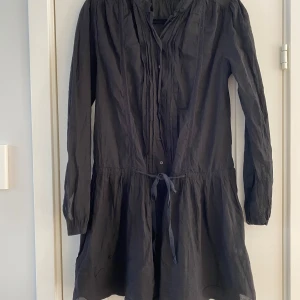 Svart kort klänning By Malene Birger - Snygg svart klänning från By Malene Birger med knyt i midjan och långa ärmar. Modellen är kort med plisserade detaljer framtill och knappar längs bröstet. Tillverkad i ett lätt och luftigt material som ger en skön känsla. Perfekt för dig som gillar stilrena och klassiska plagg.