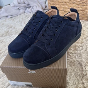 Mörkblå mocka sneakers från Louboutin - Snygga mörkblå sneakers i mocka från Louboutin med klassisk röd sula och broderad logga på hälen. Skorna har rund tå, snörning och svart yttersula. Insidan är i beige skinn för extra komfort. Perfekta för dig som gillar exklusiv streetstyle.