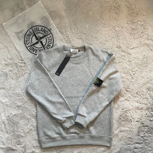 Grå sweatshirt Stone Island M - Snygg grå sweatshirt från Stone Island med klassisk rund hals och ribbade muddar. Tröjan har den ikoniska Stone Island-patchen på vänster ärm i svart, gult och grönt. Tillverkad i mjuk bomull för skön komfort och stilren look.   Skick: oanvänd 