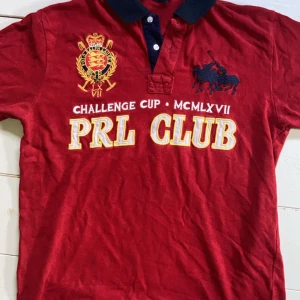 Röd PRL Club t-shirt med brodyr - Snygg röd t-shirt från PRL Club med broderade detaljer och tryck både fram och bak. Framsidan har coolt klubbmärke, text och polospelare, medan baksidan har stort nummer 3. Svart krage med knappar.