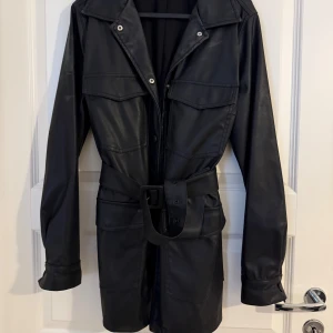 Svart skinnjacka med bälte - Snygg svart skinnjacka med bälte i midjan och stora fickor framtill. Jackan har klassisk krage, tryckknappar och långa ärmar. Perfekt för dig som vill ha en edgy och stilren look. Passar till många olika outfits och ger en cool vibe.