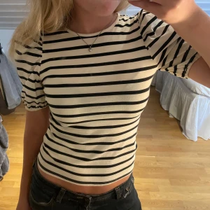 Randig puffärm topp i bomull - Säljer en snygg croppad topp med svarta och vita ränder. Toppen har puffiga korta ärmar med resår vid ärmslut och rund halsringning. Materialet är mjuk bomull och passformen är slim. Perfekt för dig som gillar trendiga detaljer och klassisk stil.