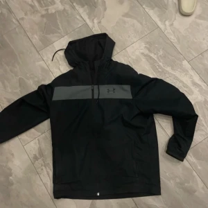 Svart vindjacka från Under Armour M - Svart vindjacka från Under Armour med grått band över bröstet och logga. Jackan har huva med snörning, dragkedja framtill och två fickor. Lätt och loose fit, perfekt för blåsiga dagar. Materialet är syntet och jackan har långa ärmar.