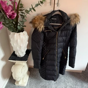 Svart lång dunjacka Rockandblue - Lång svart dunjacka från Rockandblue med snygga guldfärgade dragkedjor och quiltade detaljer på ärmar och sidor. Jackan har en stor huva med fluffig fuskpäls i bruna toner och ett emblem på ena ärmen. Perfekt för kalla dagar.