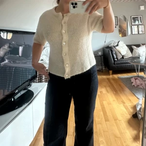 Krämvit stickad topp med knappar - Superfin krämvit stickad topp med korta ärmar och rund hals. Snygga pärlemorknappar framtill och ribbad struktur som ger en mysig och trendig vibe. Perfekt att styla med jeans eller kjol för en chill look.