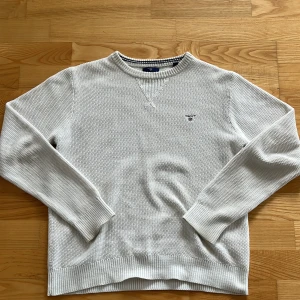 Vit stickad tröja från GANT - Säljer en stilren vit stickad tröja från GANT med rund hals och diskret logga på bröstet. Tröjan har ribbade muddar och nederkant, samt en snygg struktur i materialet. Perfekt för dig som gillar klassisk och clean stil. Tröjan har fläckar på båda handlederna som inte går bort i tvätt men dem är på undersidan så man märker inte under användning.