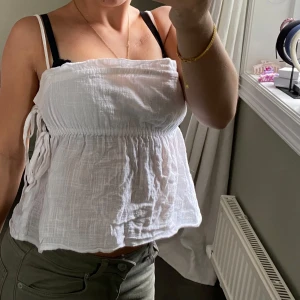 Vit linnetopp med knyt rosetter vid sidan - Säljer en vit linnetopp med smala axelband och snygga knytband i sidan samt resor för snygg look. Köpt från Nelly i storlek XS