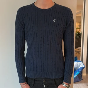 Marinblå The Original Skill Tröja - Mörkblå kabelstickad tröja från Skill med rund hals och diskret broderad S på bröstet. Tröjan har ribbade muddar och är långärmad, perfekt för lager-på-lager. Skön och klassisk stil som funkar året runt.