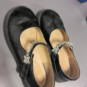 Svarta ballerinaskor med pärldetaljer - Snygga svarta ballerinaskor i lack med rund tå och dekorativa pärlbroderier på remmen. Skorna har en klassisk Mary Jane-design och är perfekta för att lyfta vilken outfit som helst med sin glansiga finish och fina detaljer.