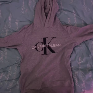 Grå hoodie från Calvin Klein Jeans - Säljer en grå hoodie från Calvin Klein med stor svart CK-logga och text på bröstet. Tröjan har huva och långa ärmar, perfekt för en chill och avslappnad stil. 