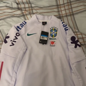Vit Brasil Nike långärmad fotbollströja - Säljer en vit långärmad Brasilien fotbollströja från Nike med CBF-emblem, grönt swoosh och flera sponsorloggor på ärmar och bröst. Tröjan har halv dragkedja vid halsen och är tillverkad i ett lätt och sportigt material.