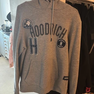 Grå hoodie från Hoodrich  - Snygg grå hoodie från Hoodrich med svart tryck och broderade patches på bröstet. Tröjan har huva med snörning och en stor magficka. Perfekt för dig som gillar streetwear och vill ha en bekväm och stilren look.
