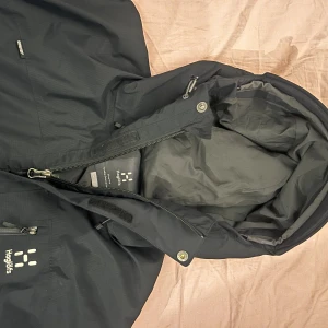 Svart vindjacka från Haglöfs med huva - Svart vindjacka från Haglöfs med justerbar huva, dragkedjor och flera fickor. Jackan är helt ny själjs pga fel storlek. Jackan är perfekt för höst/vintertid och storlek L