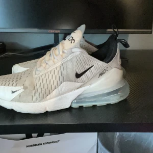 Nike Air Max 270 vita sneakers - Nike Air Max 270 sneakers i vitt med svarta detaljer och stor synlig Air-enhet i hälen. Ovandelen är i mesh med ventilerande mönster och snörning framtill. Snygg och sportig look med rund tå och platt sula, perfekt för dig som gillar streetwear.