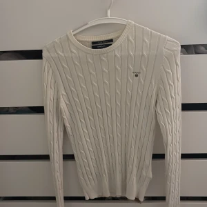 Vit Gant tröja - Vit kabelstickad tröja från Gant med rund halsringning och diskret logga på bröstet. Tröjan har lång ärm och en slim passform som sitter snyggt. Perfekt för dig som gillar klassisk och stilren look.