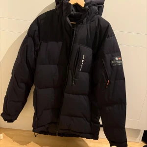 Svart Sail Racing dunjacka L - Säljer en svart dunjacka från Sail Racing, modell Antarctica Edition. Jackan har huva, flera dragkedjeförsedda fickor och tydlig logga på bröstet och ärmen. Perfekt för kalla dagar med sin rejäla fyllning och stilrena look.