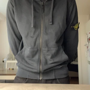 Stone Island med dragkedja - Svart hoodie från Stone Island med dragkedja framtill, huva med snörning och klassisk logga på vänster ärm. Tillverkad i mjuk bomull och har känguruficka. Perfekt för dig som gillar streetwear och vill ha en clean look.