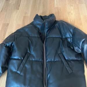 Svart pufferjacka i skinnimitation GINA TROCOT  - Säljer en svart pufferjacka med hög krage och dragkedja framtill. Jackan har en clean look med två snedställda fickor och är tillverkad i skinnimitation. Aldrig använd 💗