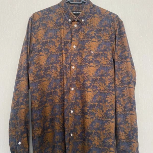 Blommig skjorta Clean Cut Copenhagen M - Snygg långärmad skjorta från Clean Cut Copenhagen i storlek M. Skjortan har ett unikt blommigt mönster i brunt och mörkblått, klassisk krage och vita knappar framtill. Perfekt för dig som vill sticka ut med en cool och stilren look.