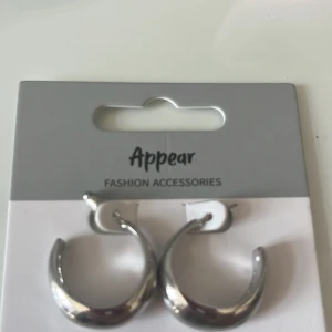 Silverfärgade hoops från Appear - Snygga och trendiga örhängen i form av chunky hoops från Appear. De är i en blank silverfärg och har en rundad, lite tjockare design som ger ett coolt intryck. Perfekta för att lyfta vilken outfit som helst. Aldrig använd