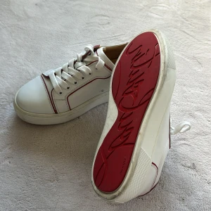 Vita sneakers från Christian Louboutin - Säljer ett par vita sneakers från Christian Louboutin med röda detaljer och ikonisk röd sula. Skorna har snörning, rund tå och är tillverkade i skinn. Snygg kontrast med röda kanter och klassisk logga på sulan. Endast använd 2 gånger. Kvitto skickas på begäran. 
