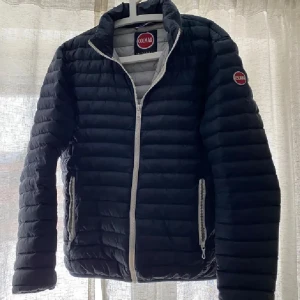 Svart pufferjacka från Colmar - Snygg svart pufferjacka från Colmar med vita dragkedjor och quiltad design. Jackan har hög krage, två fickor med dragkedja och Colmar-logga på bröstet och ärmen. Perfekt för kyliga dagar och enkel att matcha med det mesta.