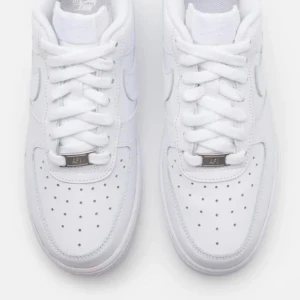 Nike Air Force 1 vita sneakers - Klassiska Nike Air Force 1 sneakers i helvitt skinn med perforerad tå, vit sula och snörning. Swoosh-logga på sidan och metallbricka med 'AF1' på snörningen. Perfekta för dig som gillar en clean och tidlös look.