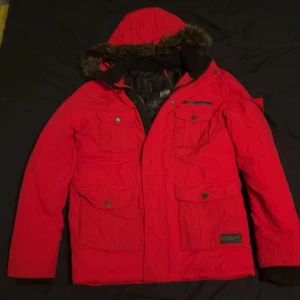 Röd parkas från Jack & Jones med fuskpäls - Röd parkas från Jack & Jones med huva och fuskpäls, flera stora fickor med tryckknappar och dragkedja framtill. Jackan är fodrad och perfekt för kalla dagar. Snygg och funktionell design som sticker ut.