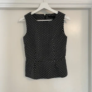 Svart prickig ärmlös blus - Snygg svart blus med vita prickar och ärmlös design. Toppen har rund halsringning och lätt figursydd passform som ger en klassisk men trendig look. Perfekt att styla med jeans eller kjol för en cool vibe.