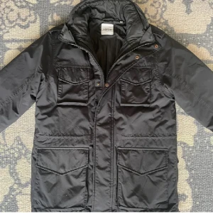 Field jacket Aspesi - Svart field jacket från Aspesi med quiltat innerfoder och flera stora fickor framtill. Jackan har dragkedja och tryckknappar. Jackans innerfoder går lätt att sätta på vilket gör den perfekt för kyliga dagar med sin stilrena look och praktiska detaljer.
