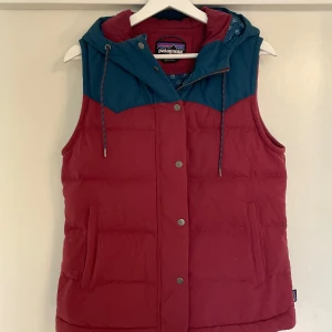 Röd och blå dunväst från Patagonia - Snygg dunväst från Patagonia i rött med blå axelparti och huva. Västen har dragkedja och tryckknappar framtill samt två fickor. Perfekt för lager-på-lager och chill utomhus. Klassisk streetstil med sportig vibe.
