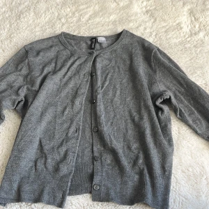 Grå kofta från H&M Divided, stl S - Grå kofta från H&M Divided i storlek S. Klassisk modell med rund halsringning och knappar framtill. Mjuk och tunn, perfekt att slänga över en t-shirt eller linne. Ribbstickade muddar vid ärmslut och nederkant.