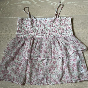 Blommig volangtop med smock och axelband - Supersöt blommig topp med smala axelband och smock upptill. Toppen har volanger och ett blommönster i rosa och vit + lite grönt. Man kan ta bort axelbanden!
